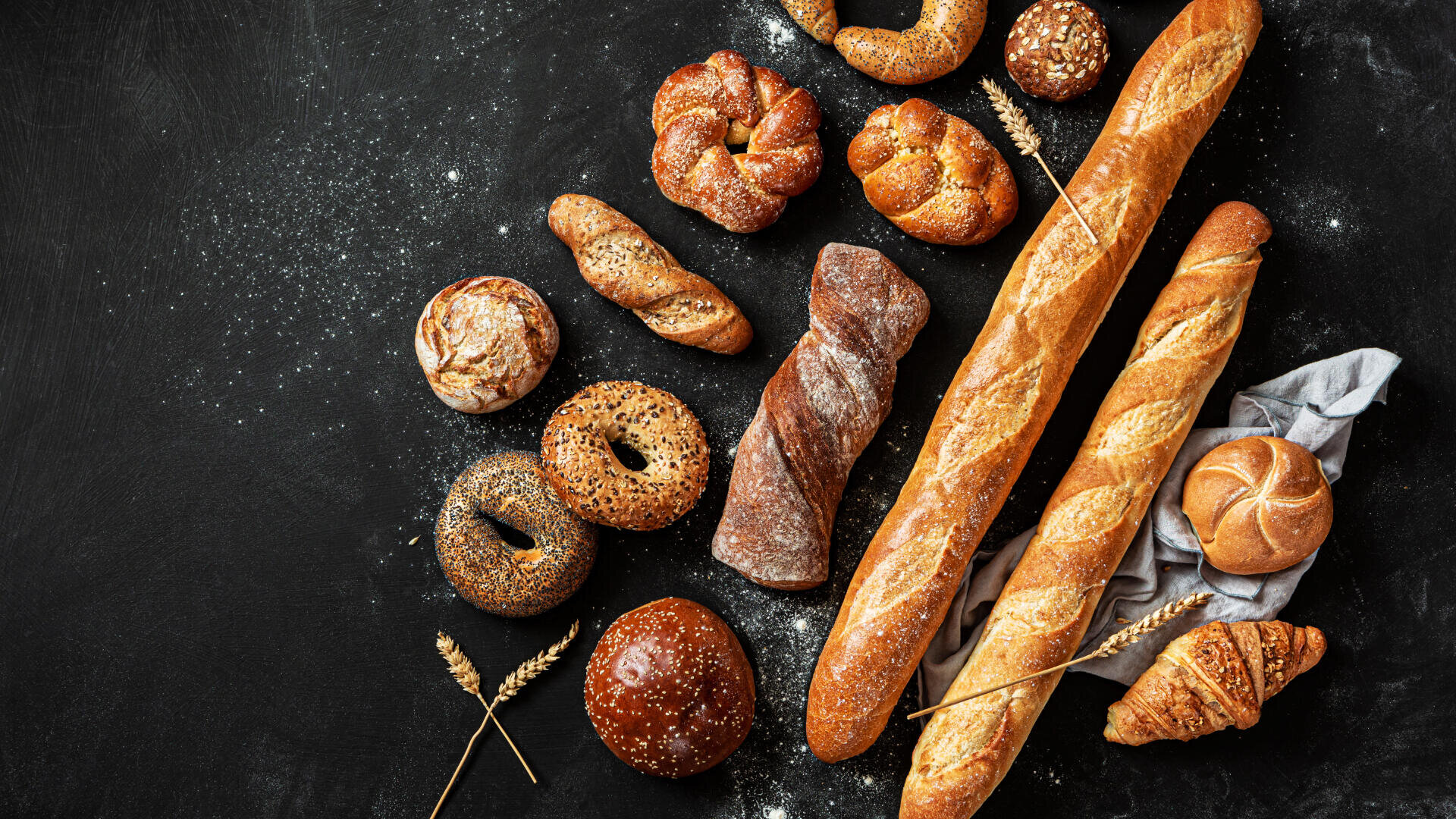 Verschiedene handwerklich hergestellte Brote und Gebäcke, darunter Baguettes, Brötchen, Brezeln, Bagels und ein Croissant, angeordnet auf einer dunklen Fläche mit Mehl und Weizenhalmen.