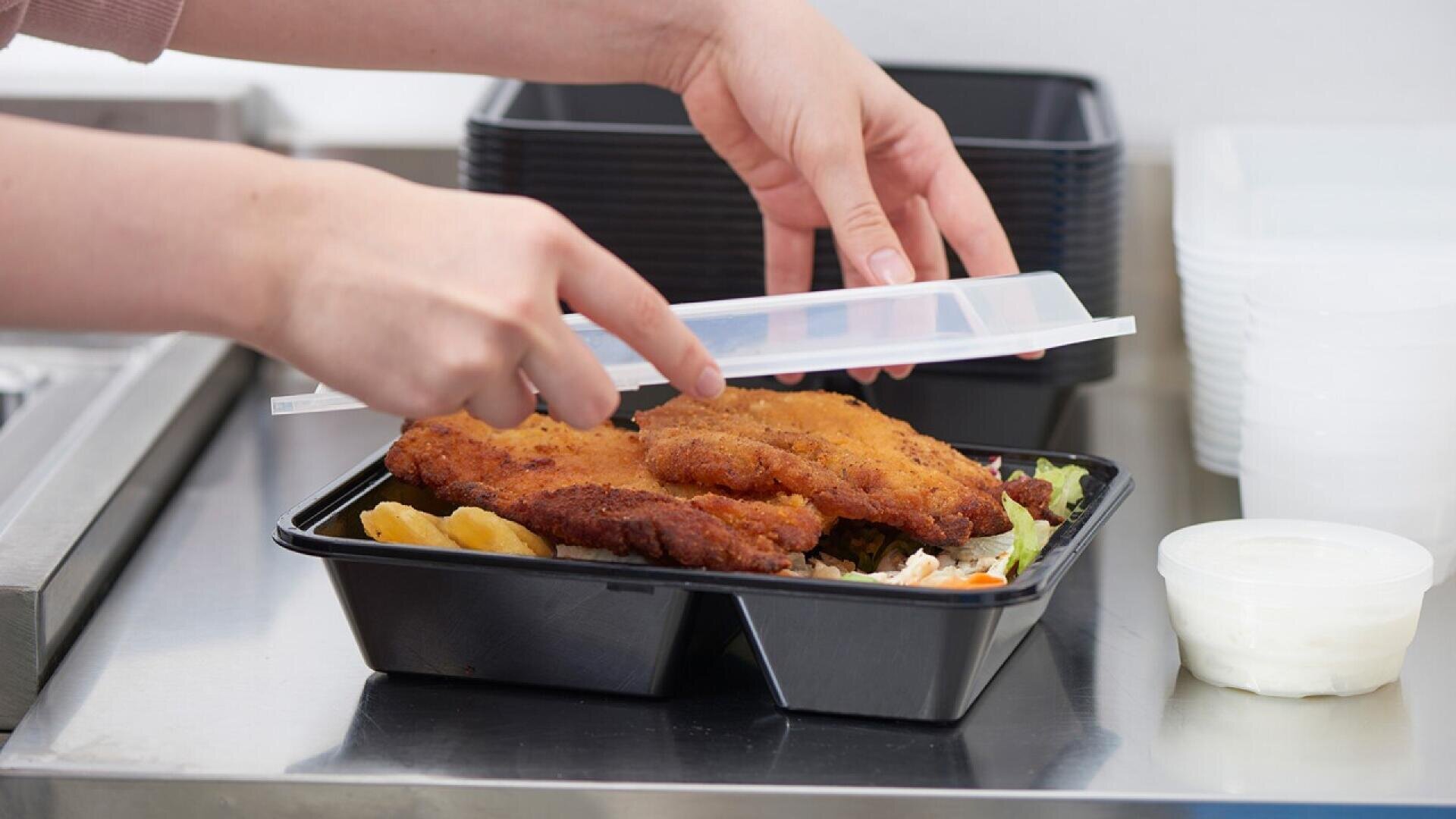 A persons hands are closing the lid on a black plastic food container filled with breaded cutlets, fries, and salad. More containers and a small sauce cup are on the counter nearby.