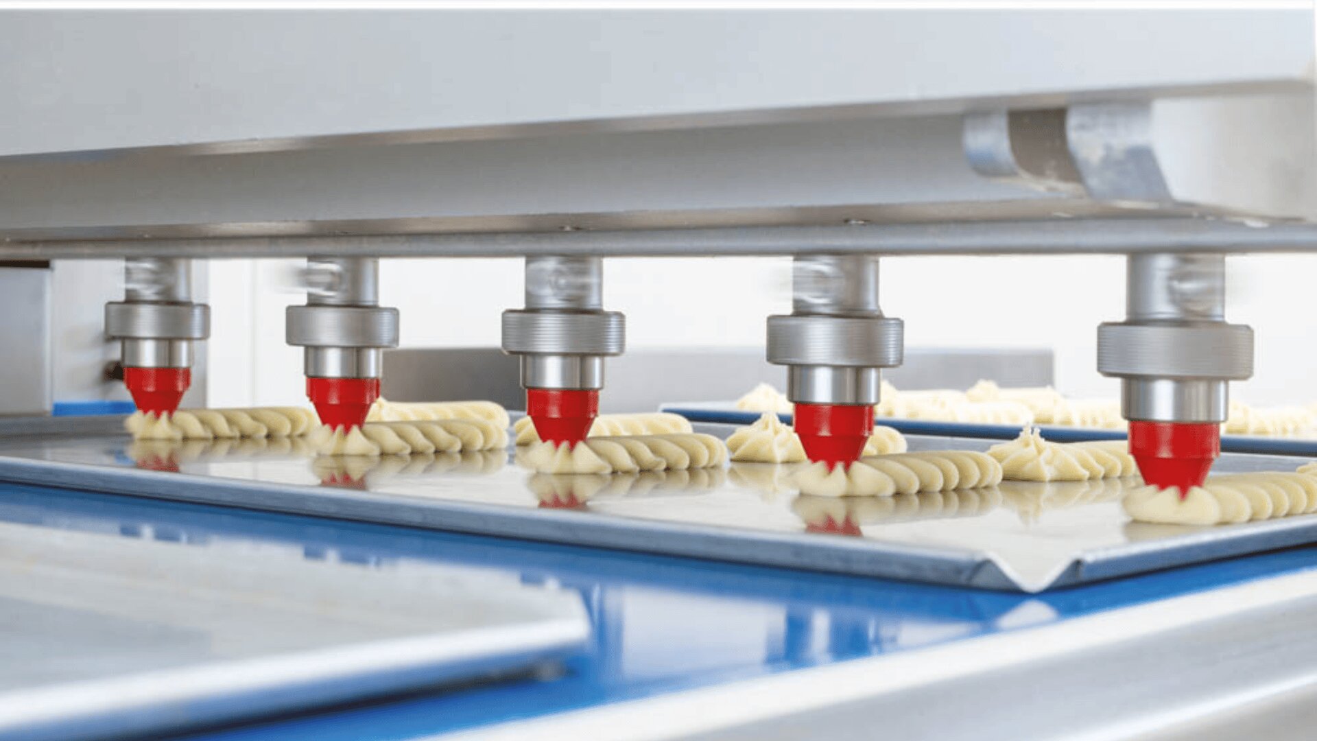 A close-up of an industrial machine with five nozzles piping dough onto metal trays, forming uniform rows of pastries or cookies in a food production facility.