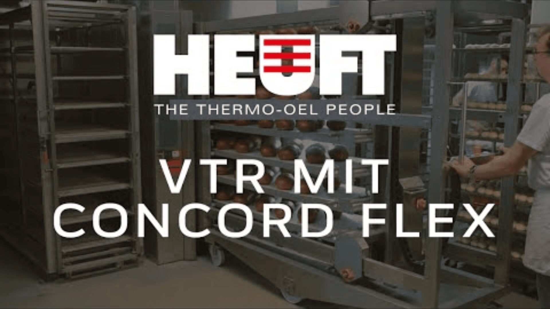 A person moves a rack of baked goods in an industrial kitchen. Overlaid text reads, HEUFT THE THERMO-OEL PEOPLE VTR MIT CONCORD FLEX. Metal racks and trays are visible in the background.