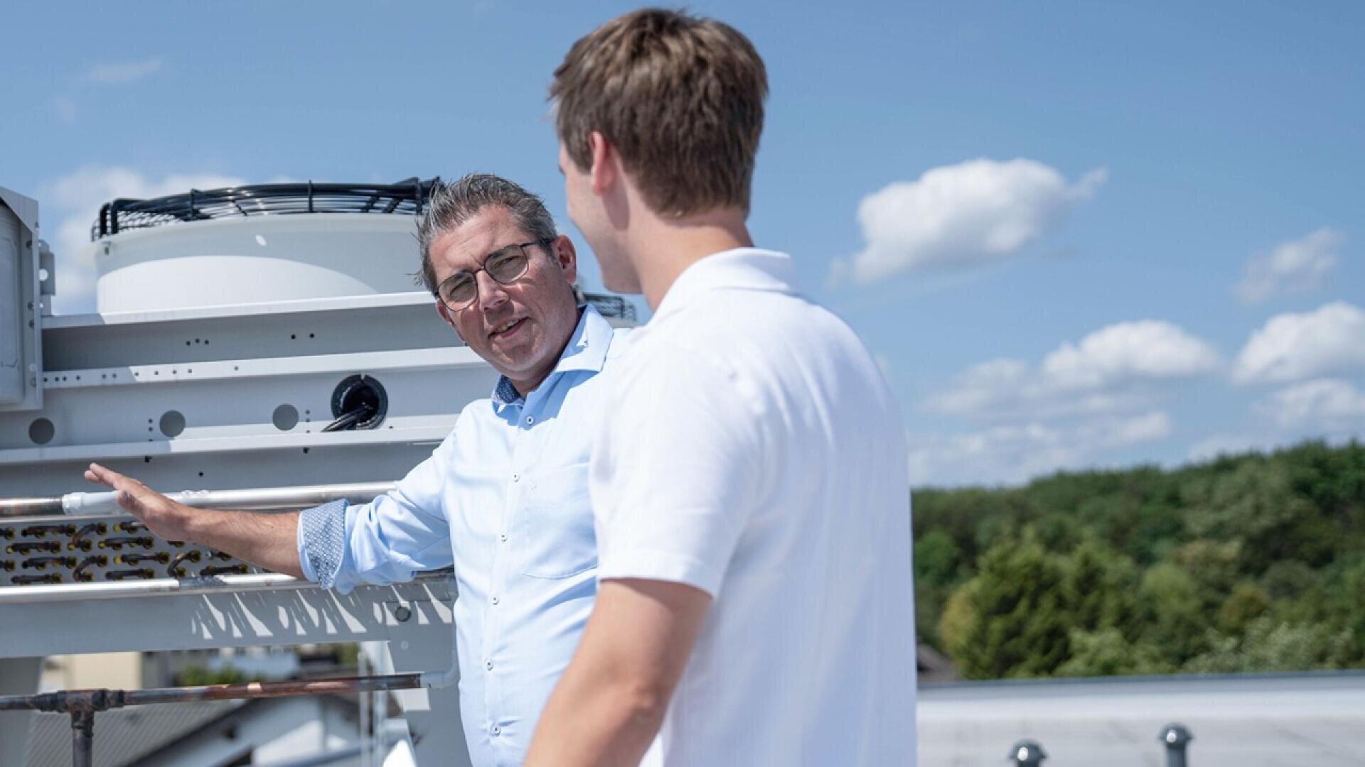 Two men stand on a rooftop near industrial equipment, having a conversation. One man points toward the equipment. Trees and a blue sky with clouds are visible in the background.
