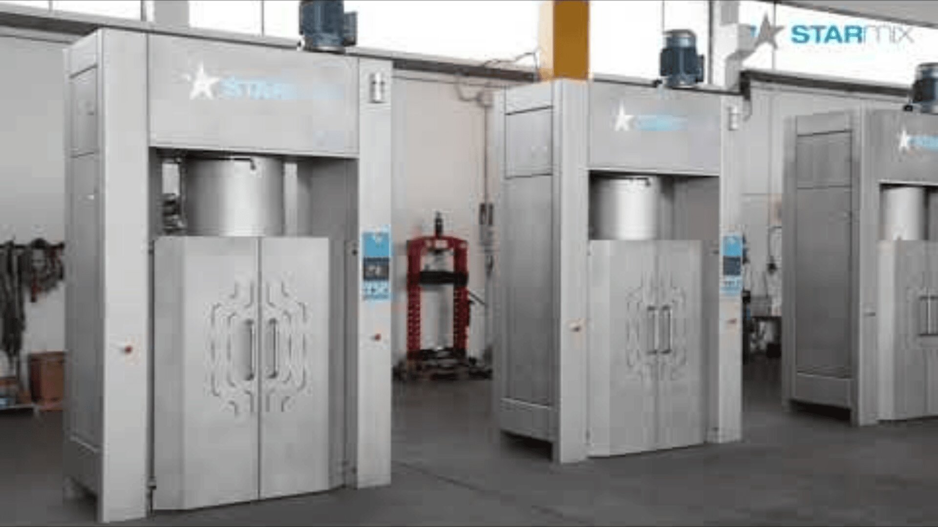 Three large industrial metal machines with closed doors stand side by side in a factory setting, with various equipment and tools visible in the background.