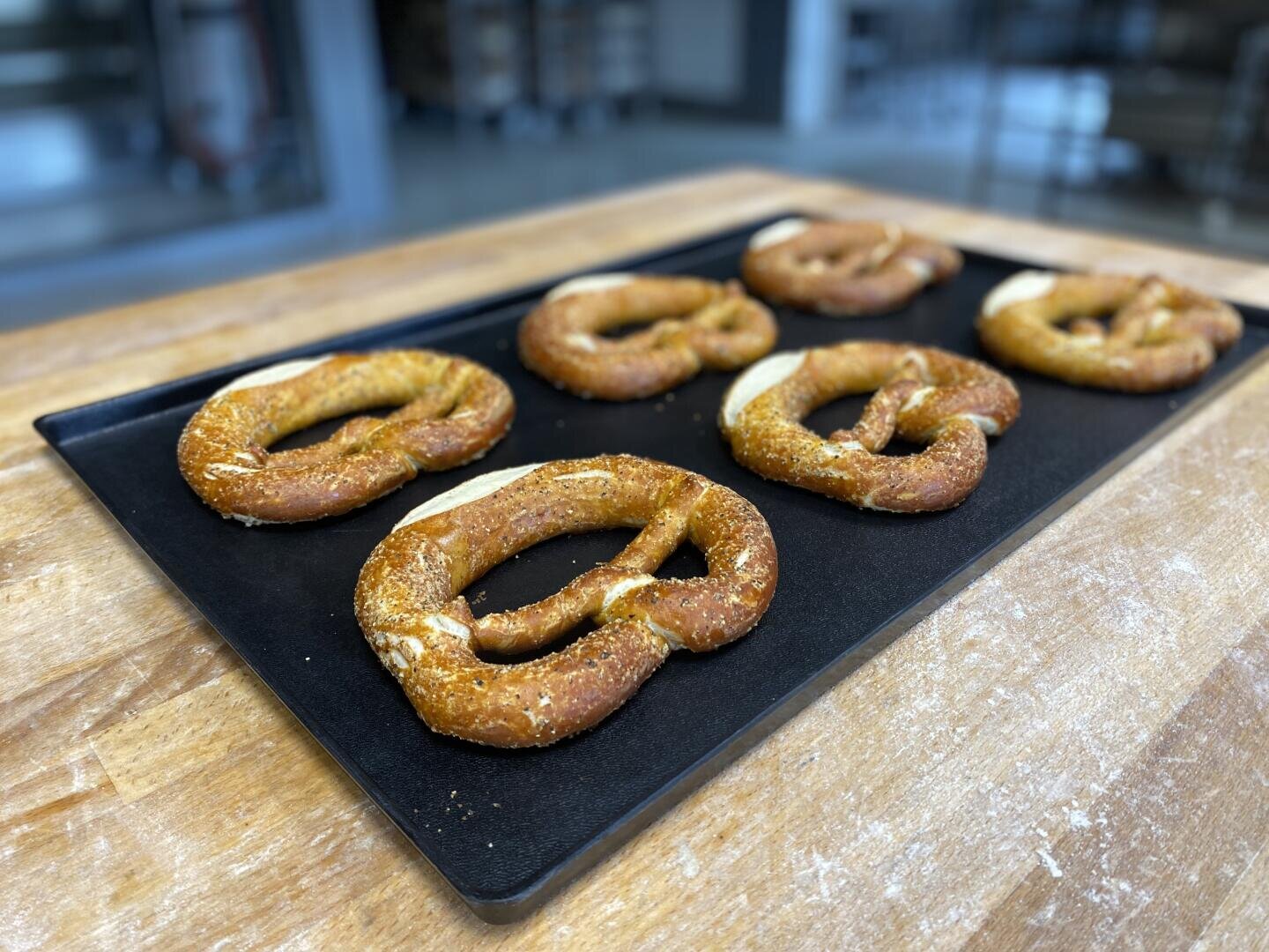 Six freshly baked soft pretzels covered in salt are arranged on a black baking tray atop a wooden counter in a kitchen or bakery setting.