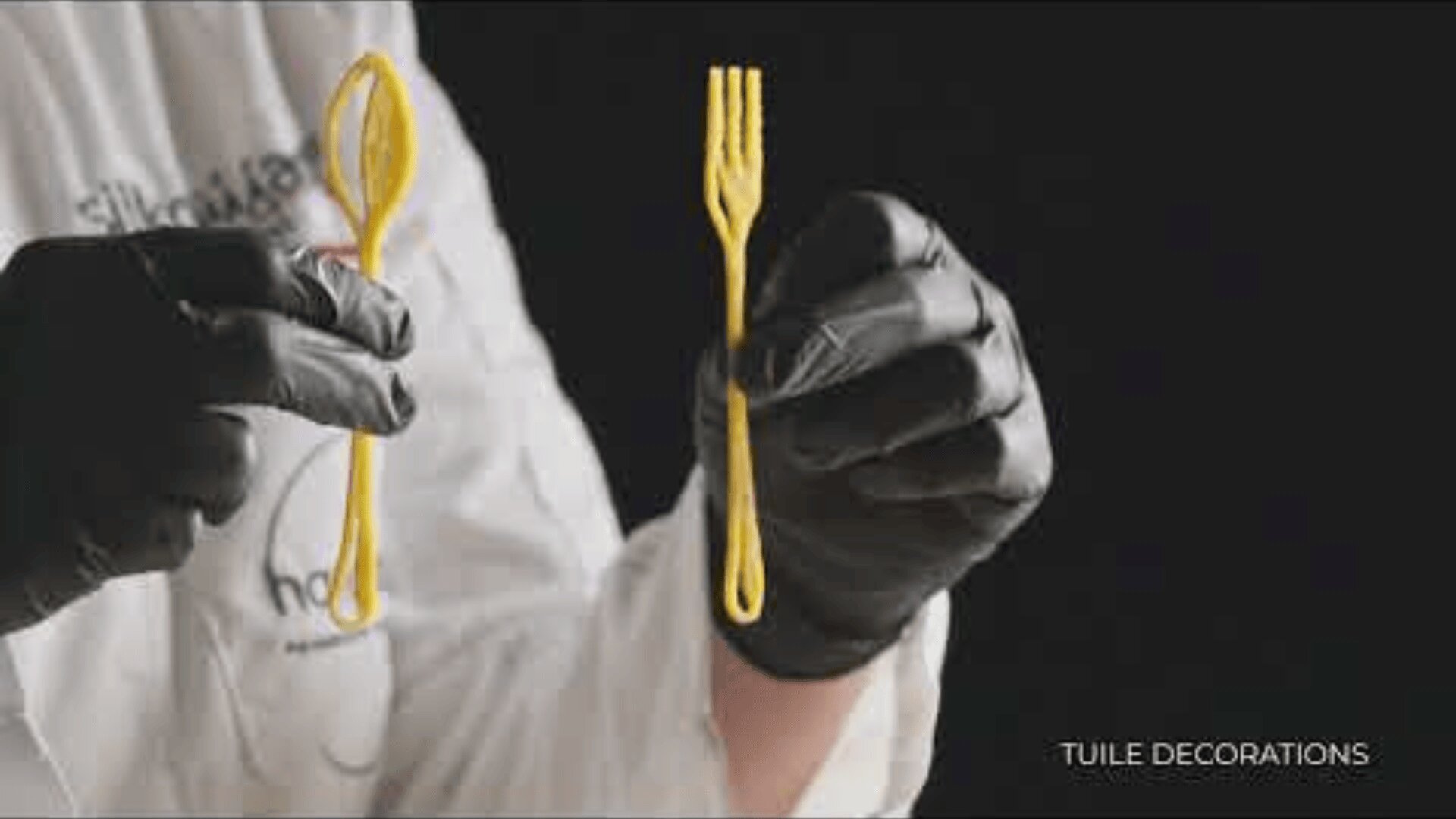 A person wearing black gloves holds a delicate, yellow, edible spoon and fork, shaped like utensils, against a dark background. The words TUILE DECORATIONS appear in the bottom right corner.