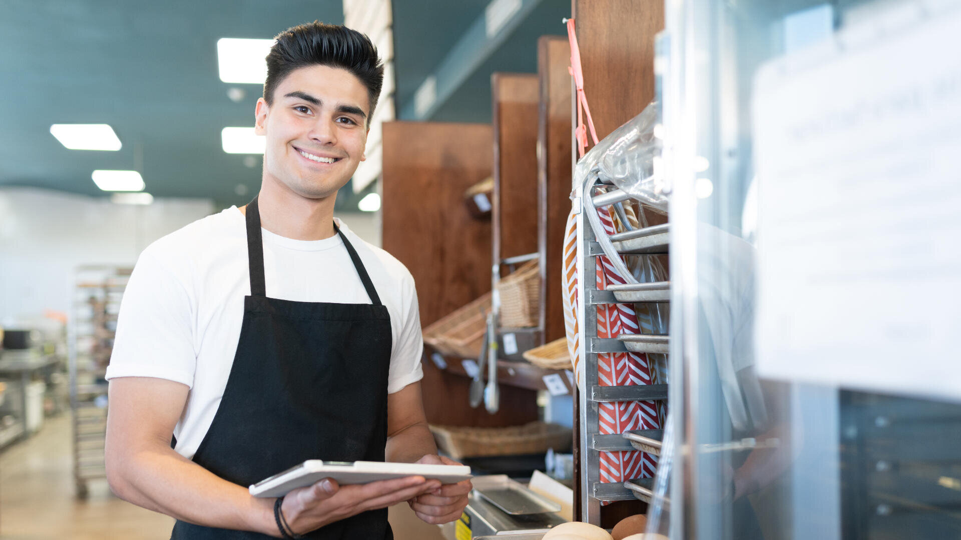 Ein junger Mann mit schwarzer Schürze und weißem Hemd lächelt, während er in einer Bäckerei oder einem Café ein Tablet hält. Hinter ihm stehen leere Körbe und Regale, die auf einen Einzelhandels- oder Gastronomiebetrieb hindeuten.