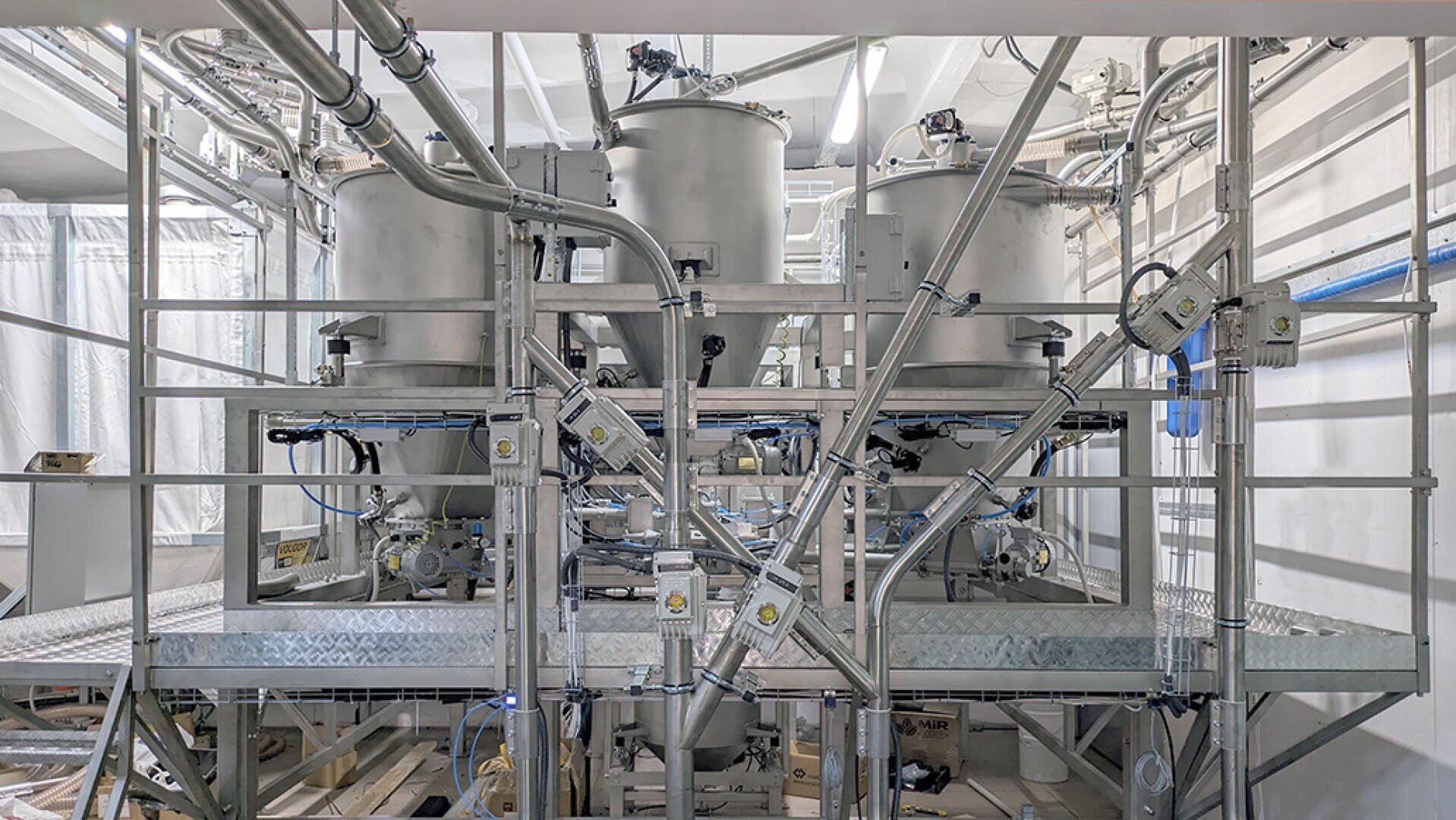 Industrial equipment featuring several large stainless steel tanks connected by multiple pipes and valves, all mounted on a metal platform inside a clean, well-lit facility.