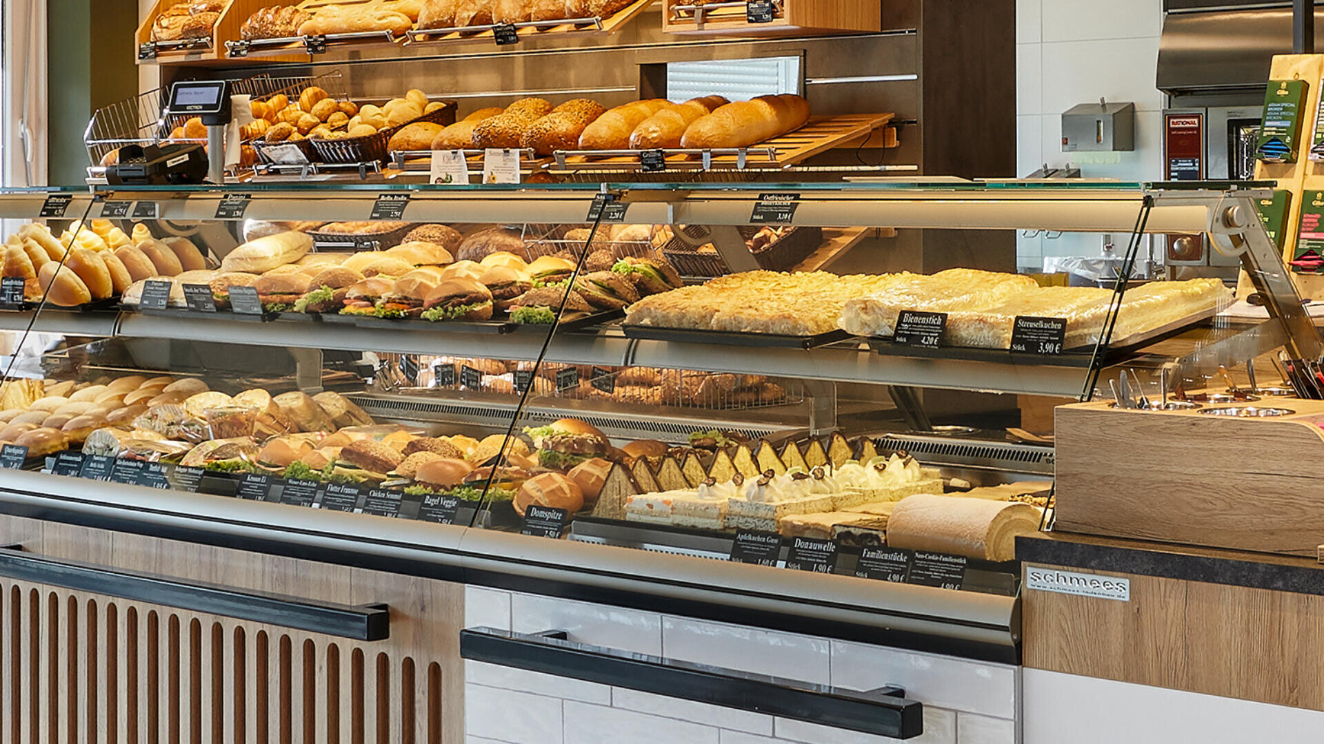 Eine Bäckereivitrine, gefüllt mit verschiedenen Brotsorten, Sandwiches, Gebäck und Desserts. Brote und Brötchen sind in den Regalen über der Glasvitrine angeordnet, während im Inneren Kuchen und gebackene Leckereien ordentlich präsentiert werden.
