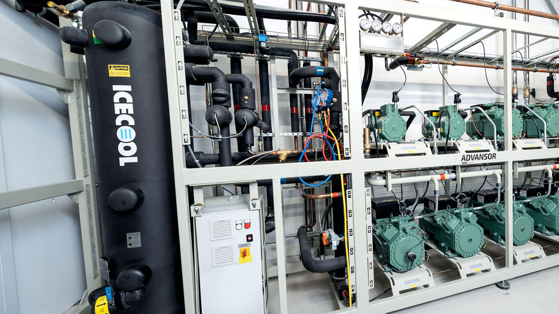 A commercial refrigeration system with multiple green compressors, labeled components, gauges, pipes, wiring, and a large black vertical tank labeled ICECOOL inside an industrial facility.