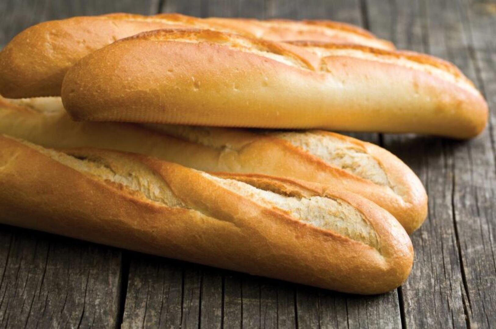 Four golden-brown baguettes are stacked on a rustic wooden surface. The bread has a crisp crust with characteristic diagonal slashes on top.