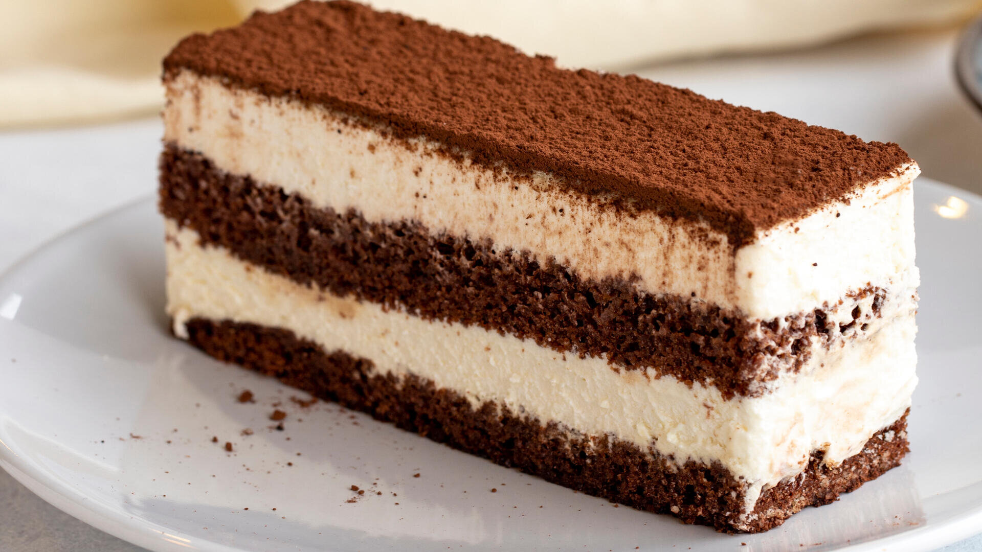Ein Stück geschichteter Schokoladen-Sahne-Torte, bestäubt mit Kakaopulver, serviert auf einem weißen Teller.