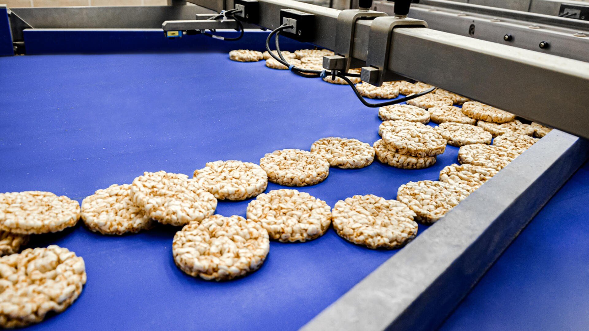 Round rice cakes travel along a blue conveyor belt in a food processing facility, passing under machinery as part of an automated production line.