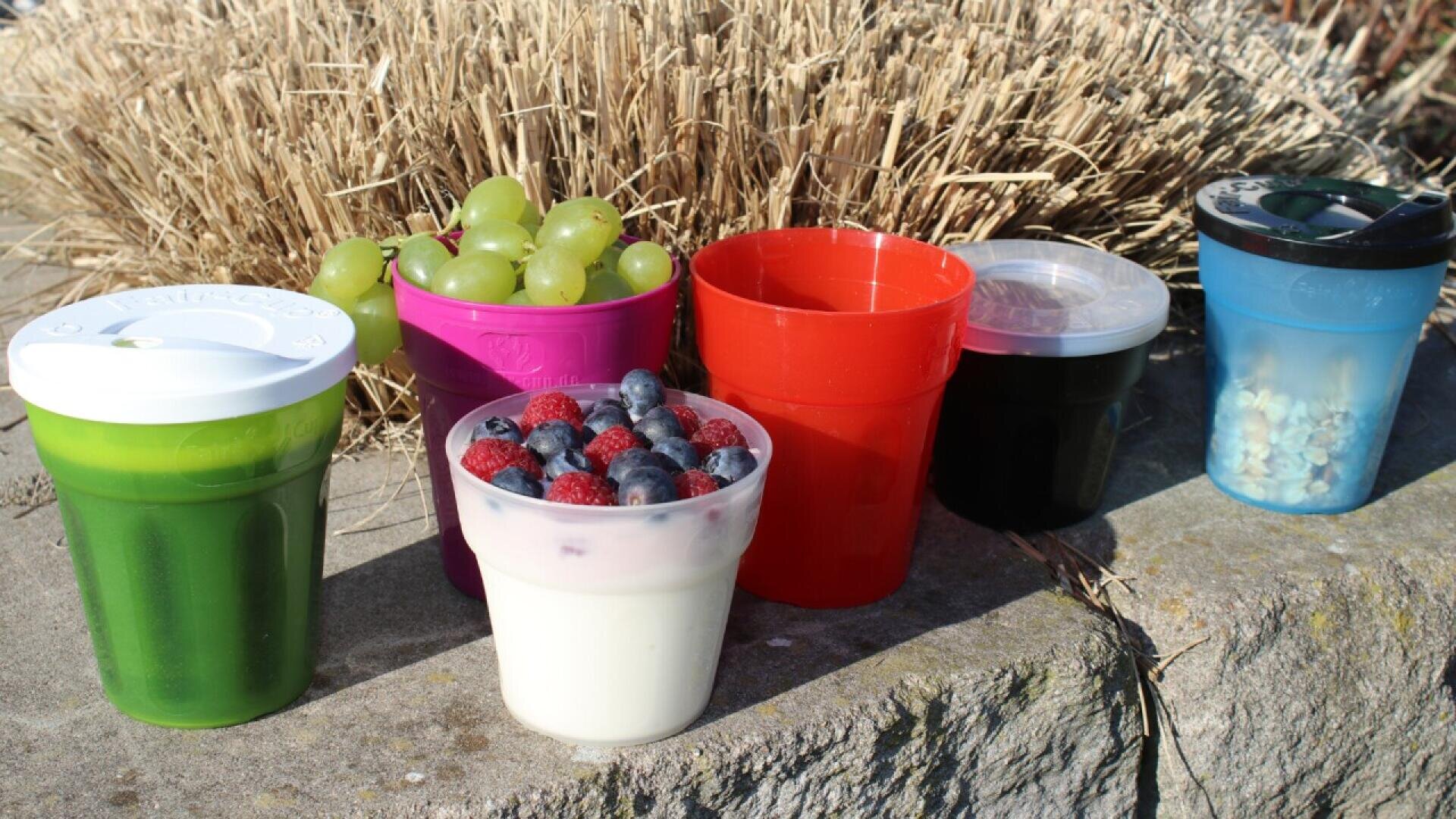 Five colorful cups are lined up on a stone surface outdoors. The cups contain green smoothie, green grapes, yogurt with berries, a dark liquid, and popcorn. Dry tall grass is in the background.