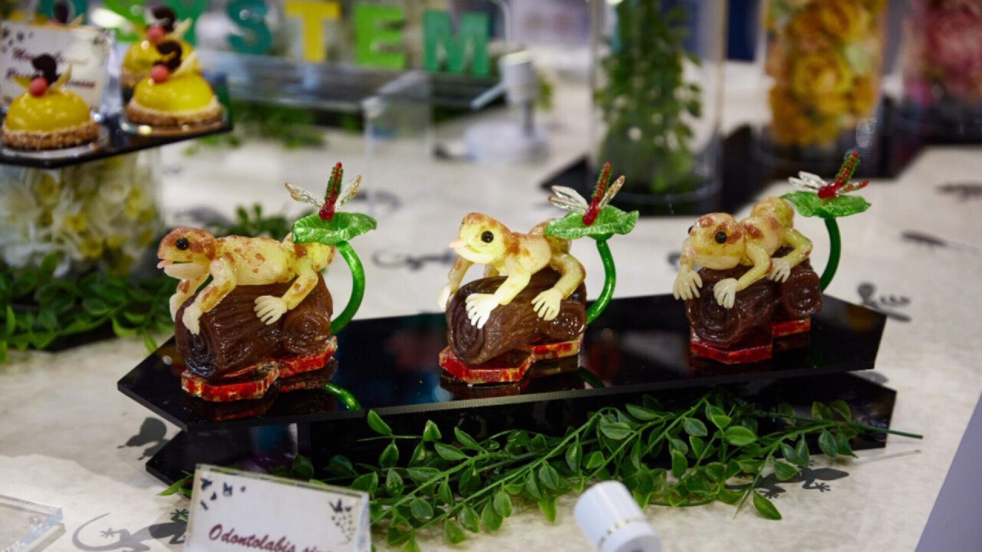 Three detailed, frog-shaped desserts sit on a glossy black tray, each perched on a chocolate log with a green candy stem and a decorative dragonfly. Green leaves decorate the base, and other colorful treats are visible in the background.