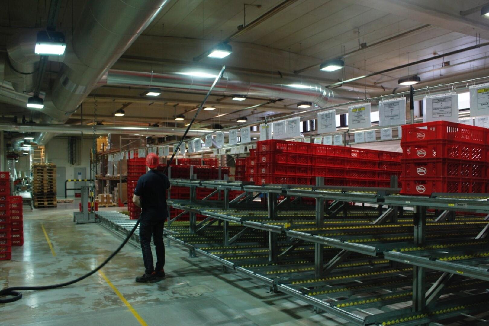 Eine Person steht in einer Lagerhalle mit roten Plastikkisten auf Metallregalen und hält unter hellem Deckenlicht einen langen schwarzen Schlauch in der Hand. Über den Regalen sind Schilder angebracht, im Hintergrund sind leere Paletten gestapelt.