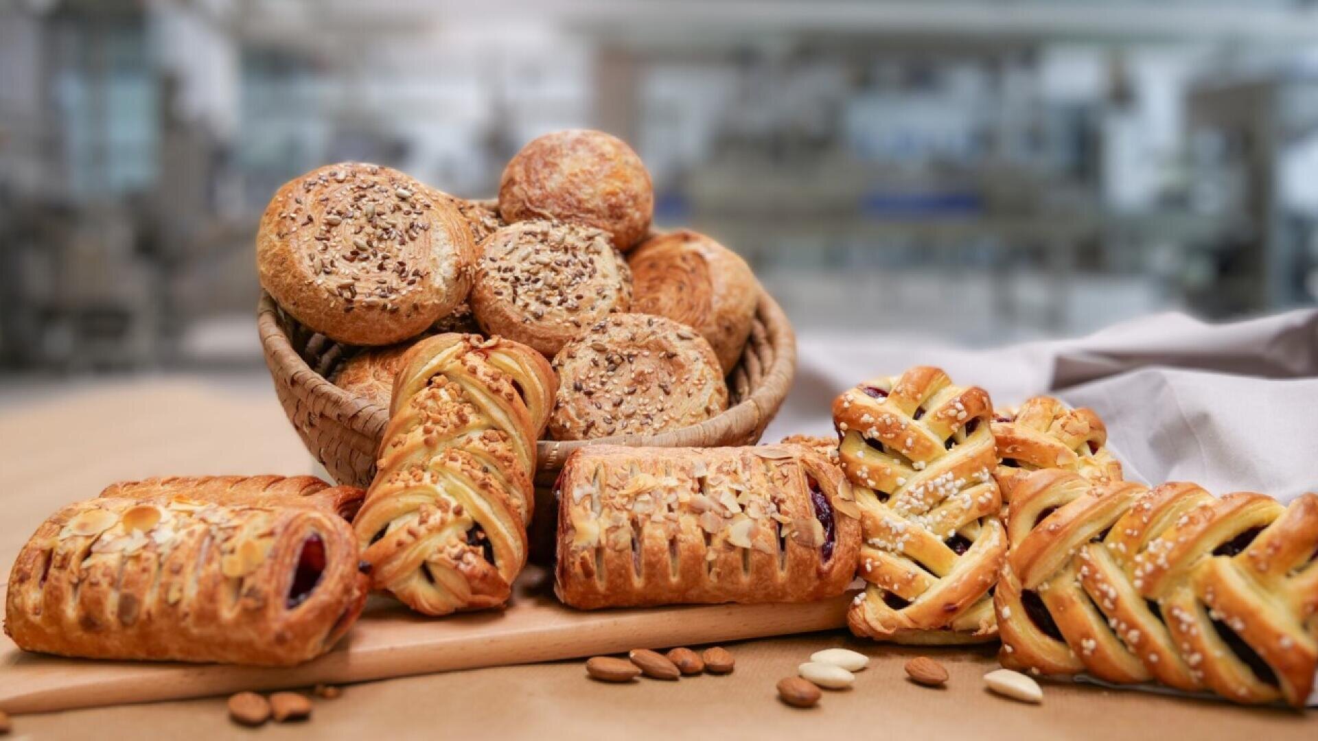 A variety of pastries, including seeded rolls and fruit danishes, are displayed on a wooden board and in a basket, with a blurred bakery kitchen in the background. Almonds and seeds are scattered in front.