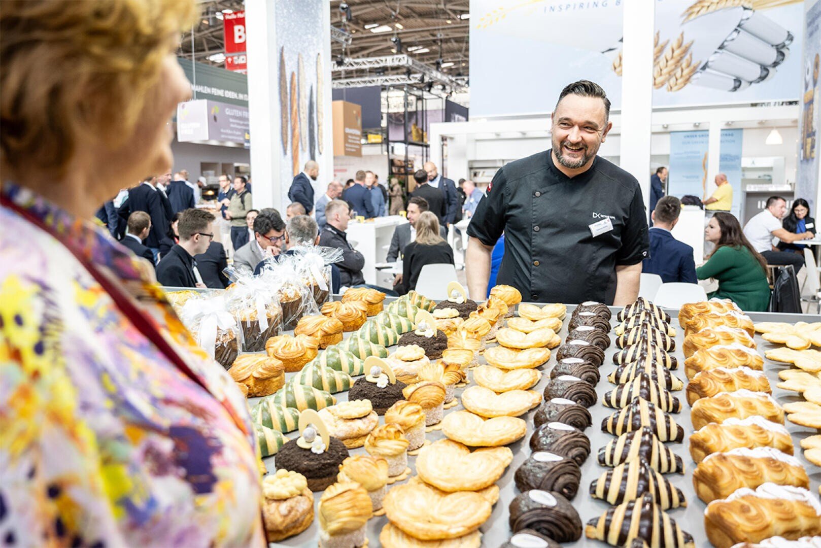 A smiling chef stands behind a display of assorted pastries at a busy indoor event, speaking to a woman in a colorful blouse. People are seated and talking in the background.