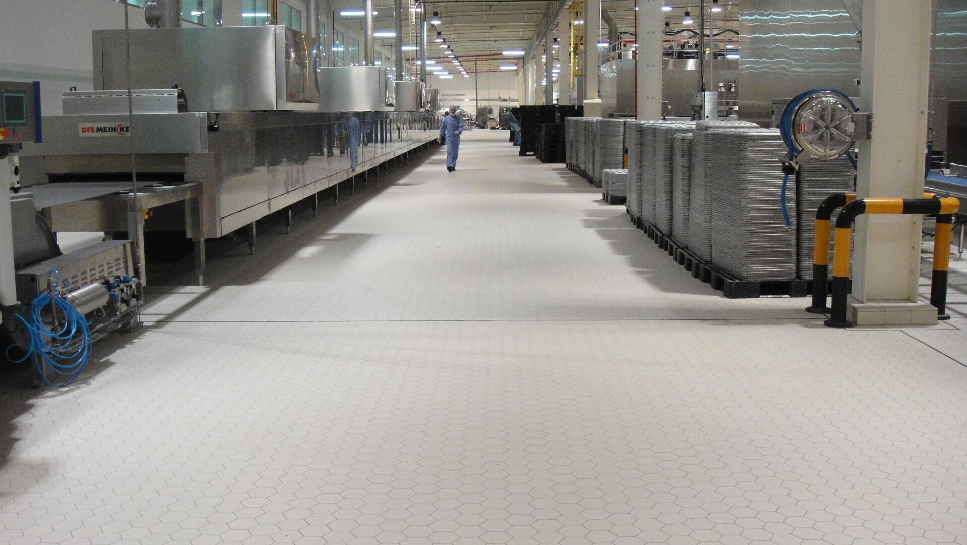 A worker in a blue uniform walks down a clean, well-lit factory floor lined with industrial machines and stacked metal trays on the right.