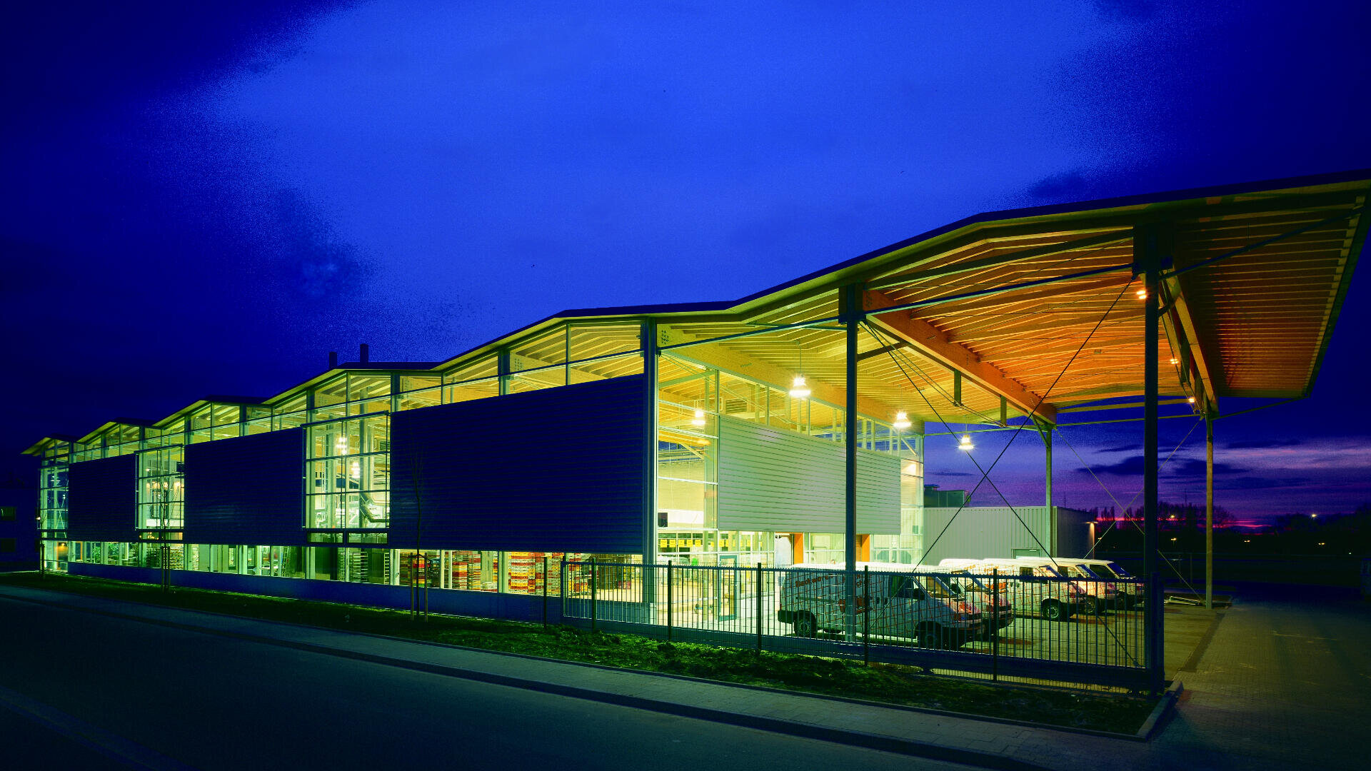 A modern, brightly lit building with large glass walls and a zigzag roof stands against a deep blue evening sky; outside, delivery vans are parked along a fence.