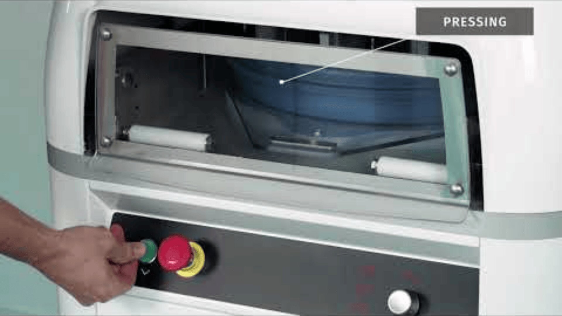 A person operates a machine by pressing a red button on a control panel. The machine has a window with the internal components and a label with the word PRESSING in the top right corner.