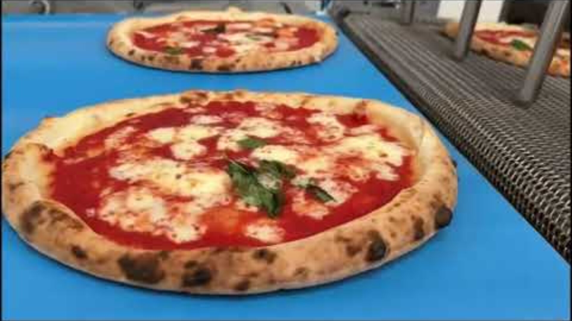 Zwei Margherita-Pizzen mit Tomatensoße, geschmolzenem Mozzarella und Basilikumblättern liegen auf einem blauen Förderband in einer Umgebung, die wie eine Großküche oder eine Pizzaproduktion aussieht.