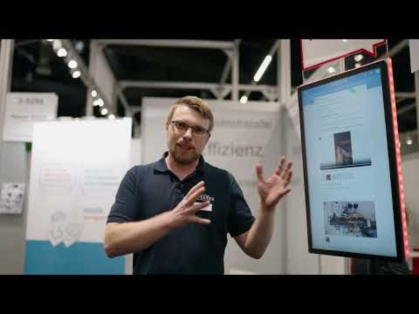 A man in glasses and a navy polo shirt gestures with his hands while standing next to a large vertical screen displaying a webpage at an indoor exhibition or trade show booth.