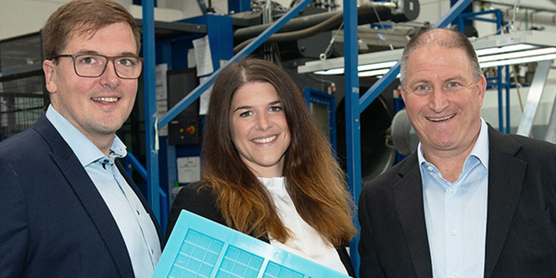 Three people in business attire stand indoors, smiling at the camera. The woman in the center holds a blue plastic tray. Industrial equipment and machinery are visible in the background.