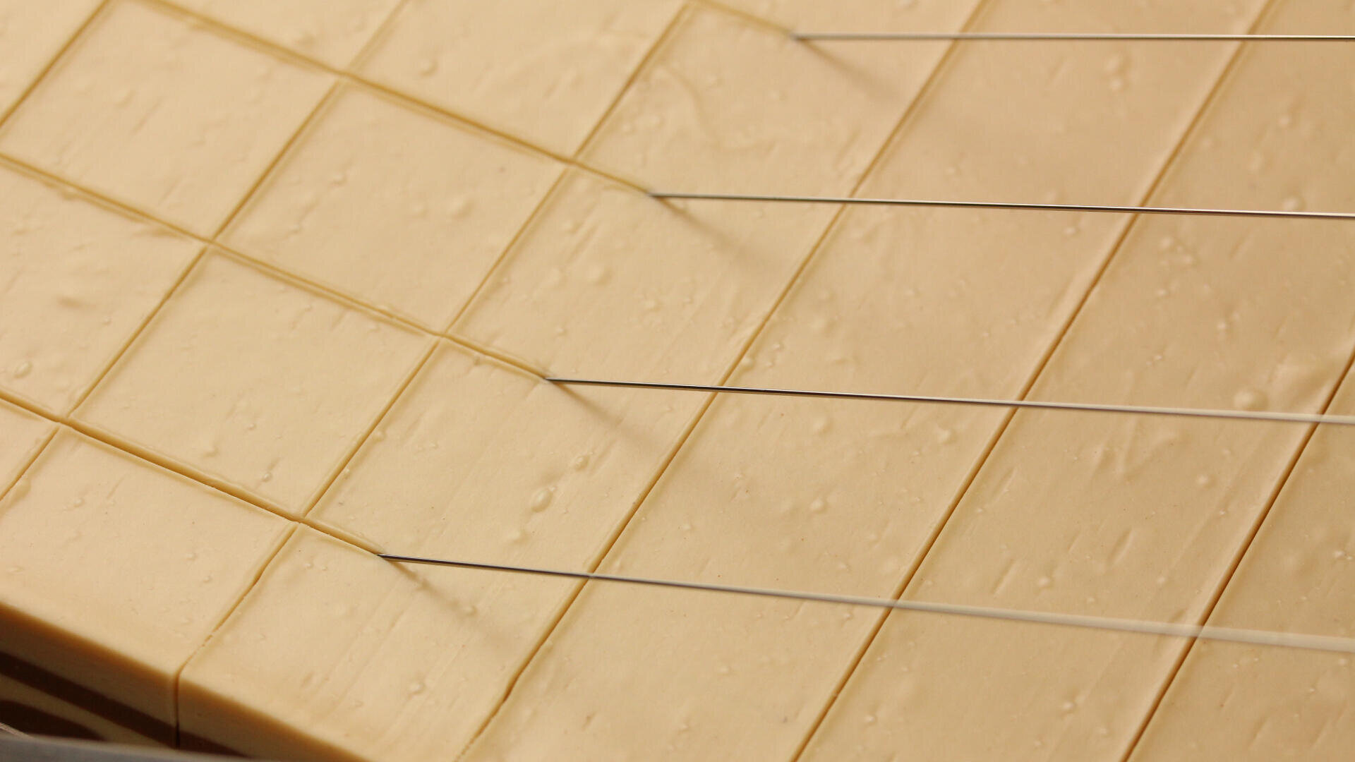 Nahaufnahme eines Käseblocks, der mit dünnen Metalldrähten an einer Käseschneidemaschine in gleichmäßige Stücke geschnitten wird. Der Käse hat eine glatte, blassgelbe Oberfläche mit ein paar kleinen Löchern.