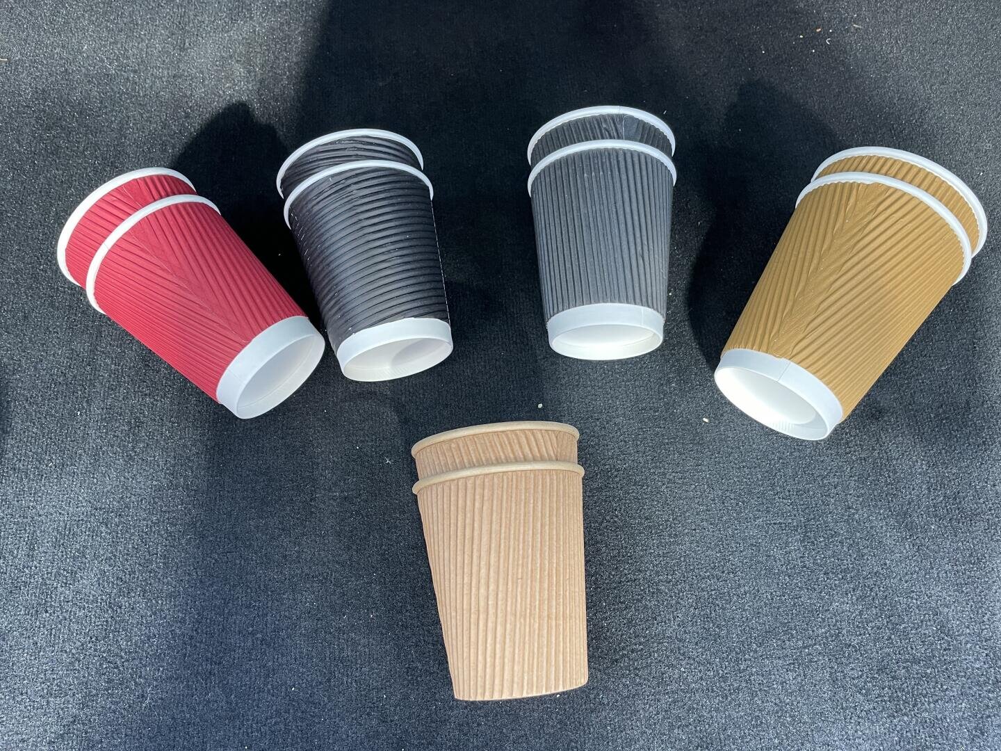 Five ribbed paper coffee cups in different colors (red, black, gray, yellow, and brown) are arranged on a dark surface, with one brown cup centered and the others fanned out above it.