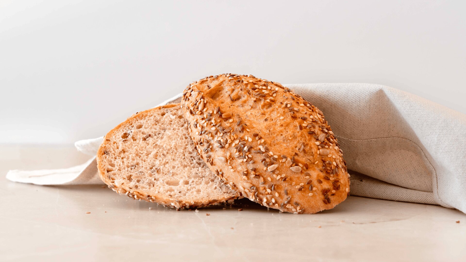 Zwei Scheiben Mehrkornbrot mit sichtbaren Körnern liegen auf einer hellen Oberfläche, teilweise mit einem beigen Tuch bedeckt, vor einem schlichten weißen Hintergrund.