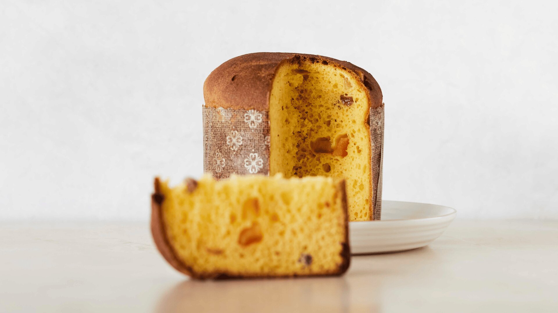 Auf einem weißen Teller liegt ein runder Laib Panettone mit goldbrauner Kruste. Eine große Scheibe ist abgeschnitten und in den Vordergrund gelegt, so dass das gelbe, luftige Innere mit Trockenfruchtstückchen zu sehen ist.