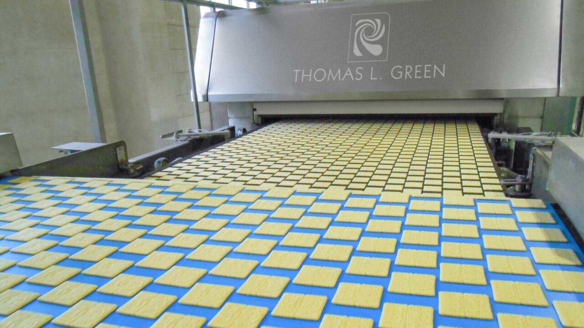 Reihen von rechteckigen Crackern laufen über ein Förderband in einen großen industriellen Backofen mit der Aufschrift "THOMAS L. GREEN". Die Kekse sind gleichmäßig verteilt und die Umgebung scheint eine Lebensmittelproduktionsstätte zu sein.