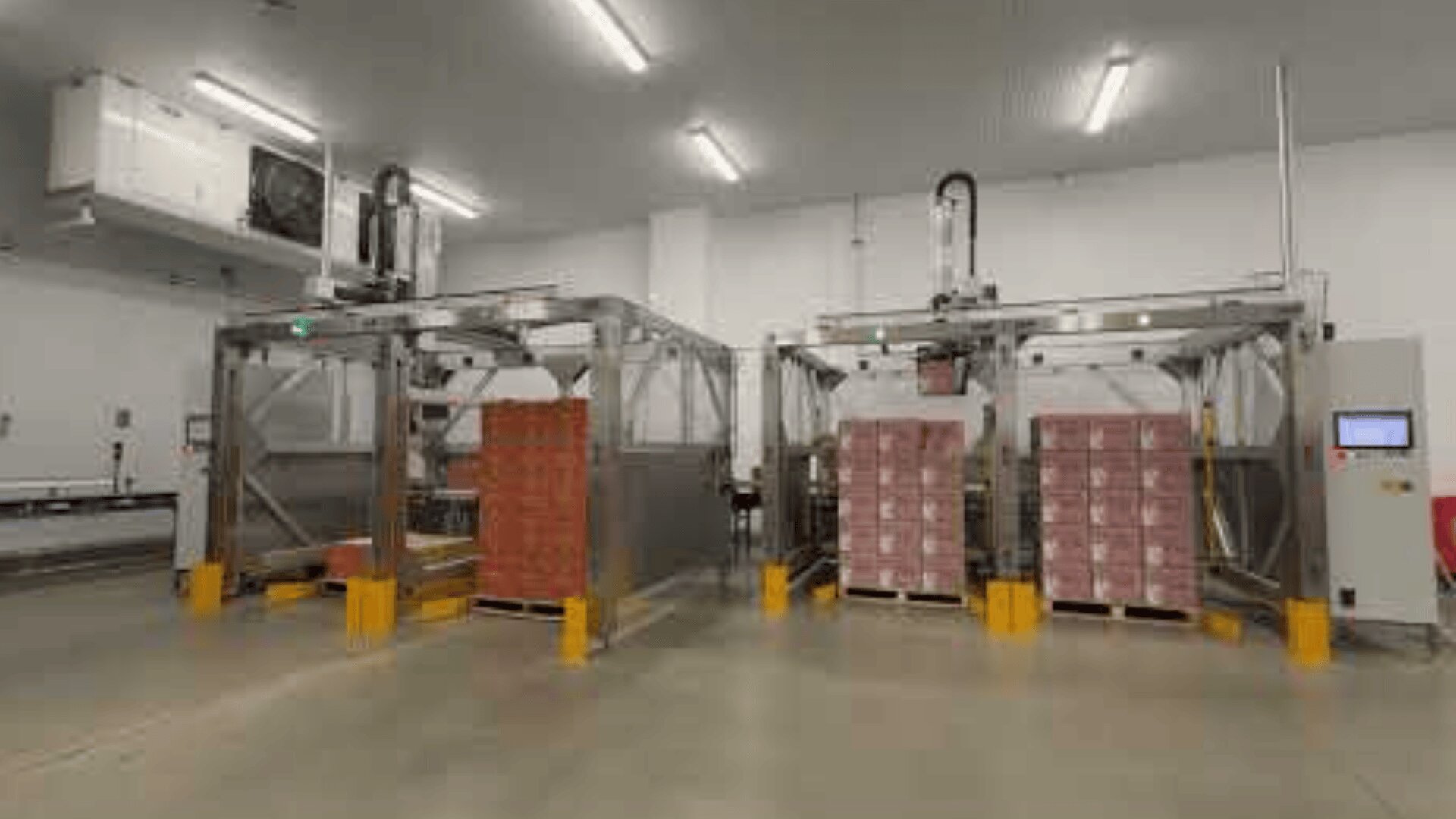 Two automated palletizing machines stack pallets of red-labeled boxes in a clean, well-lit industrial facility with white walls and a concrete floor. Conveyor belts and control panels are also visible.