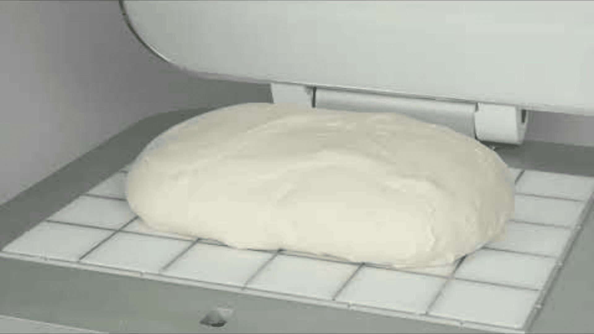 A large piece of bread dough rests on a grid-like surface under a partially opened lid of an industrial machine, possibly for kneading or pressing.