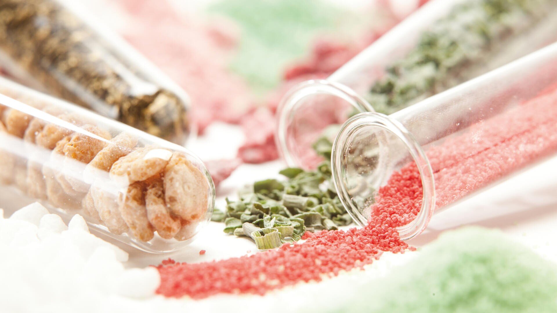 Several glass test tubes lie on a white surface, spilling colorful contents such as red, green, and white granules, as well as dried herbs and round pellets. The granules and herbs form colorful scattered piles.