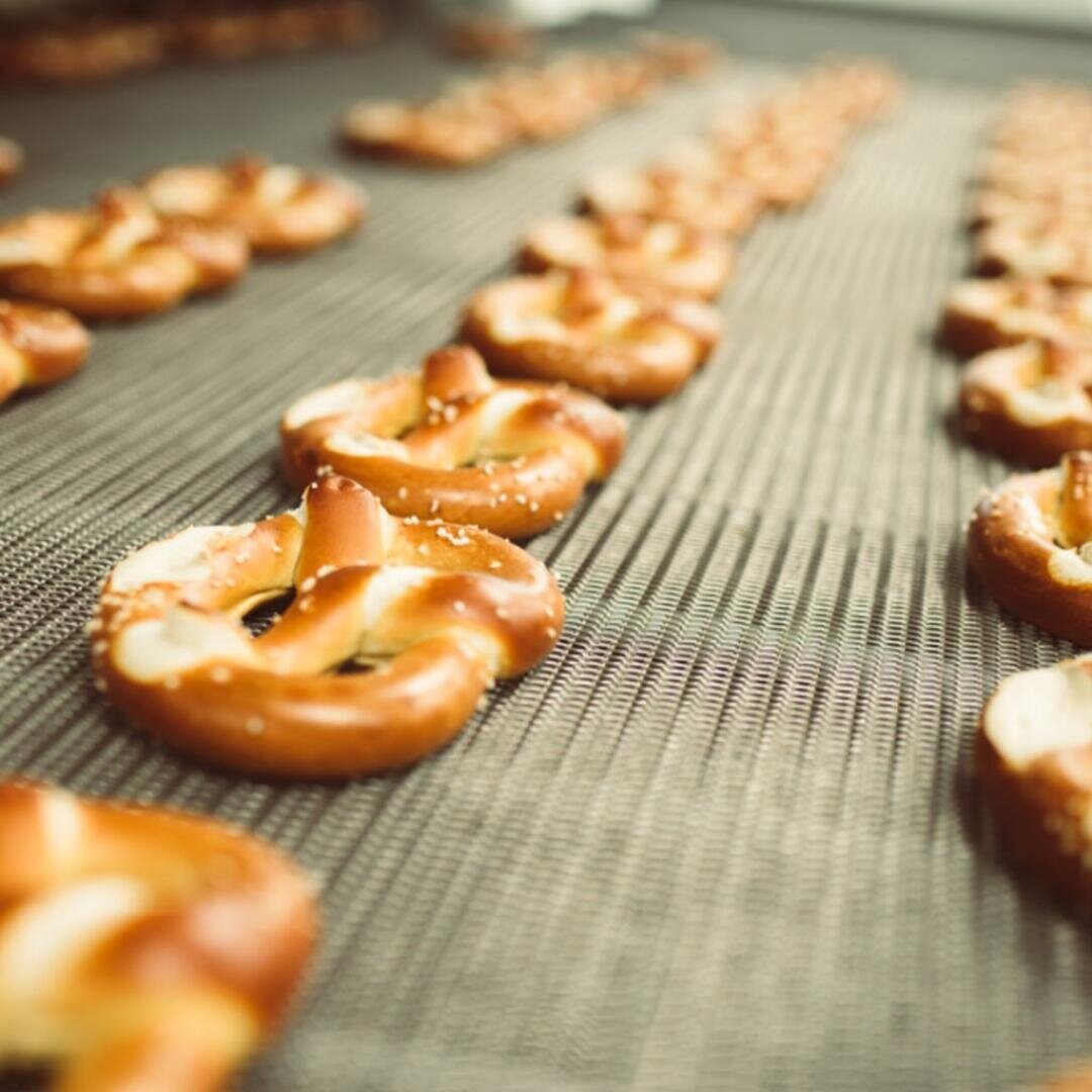 Reihen von frisch gebackenen Brezeln mit gestreutem Salz sind auf einem Förderband aufgereiht, wahrscheinlich in einer Bäckerei oder Fabrik. Das Bild konzentriert sich auf die Brezeln im Vordergrund.
