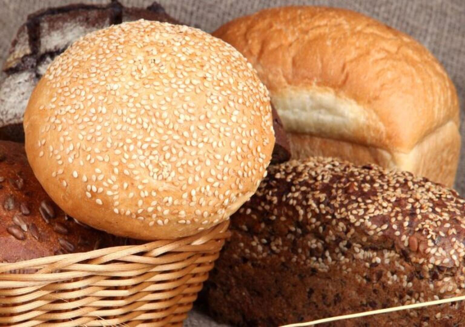 Assorted breads including a sesame seed bun, a loaf of white bread, and various seeded whole grain loaves, arranged together with part of a wicker basket visible in the foreground.