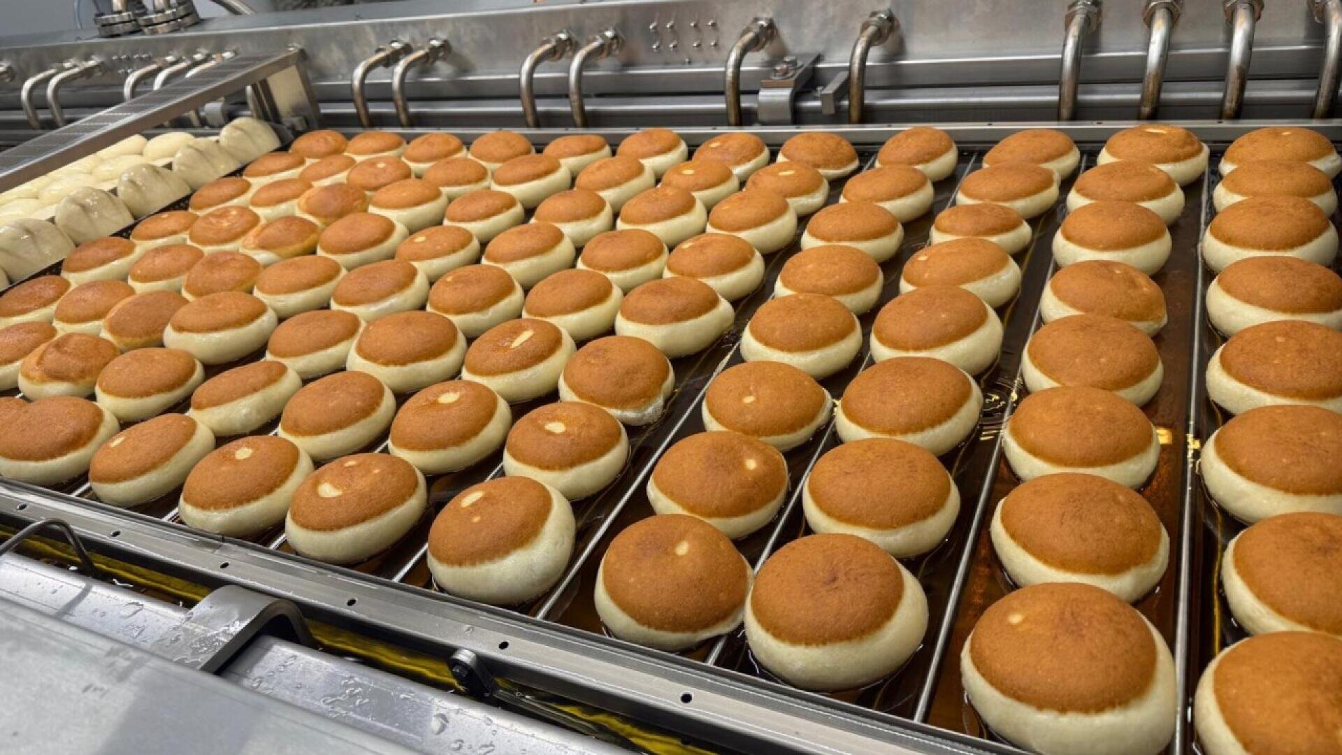 Rows of round, golden-brown buns move along a metal conveyor belt in a commercial bakery, with metal pipes above, likely part of an automated baking or cooling process.