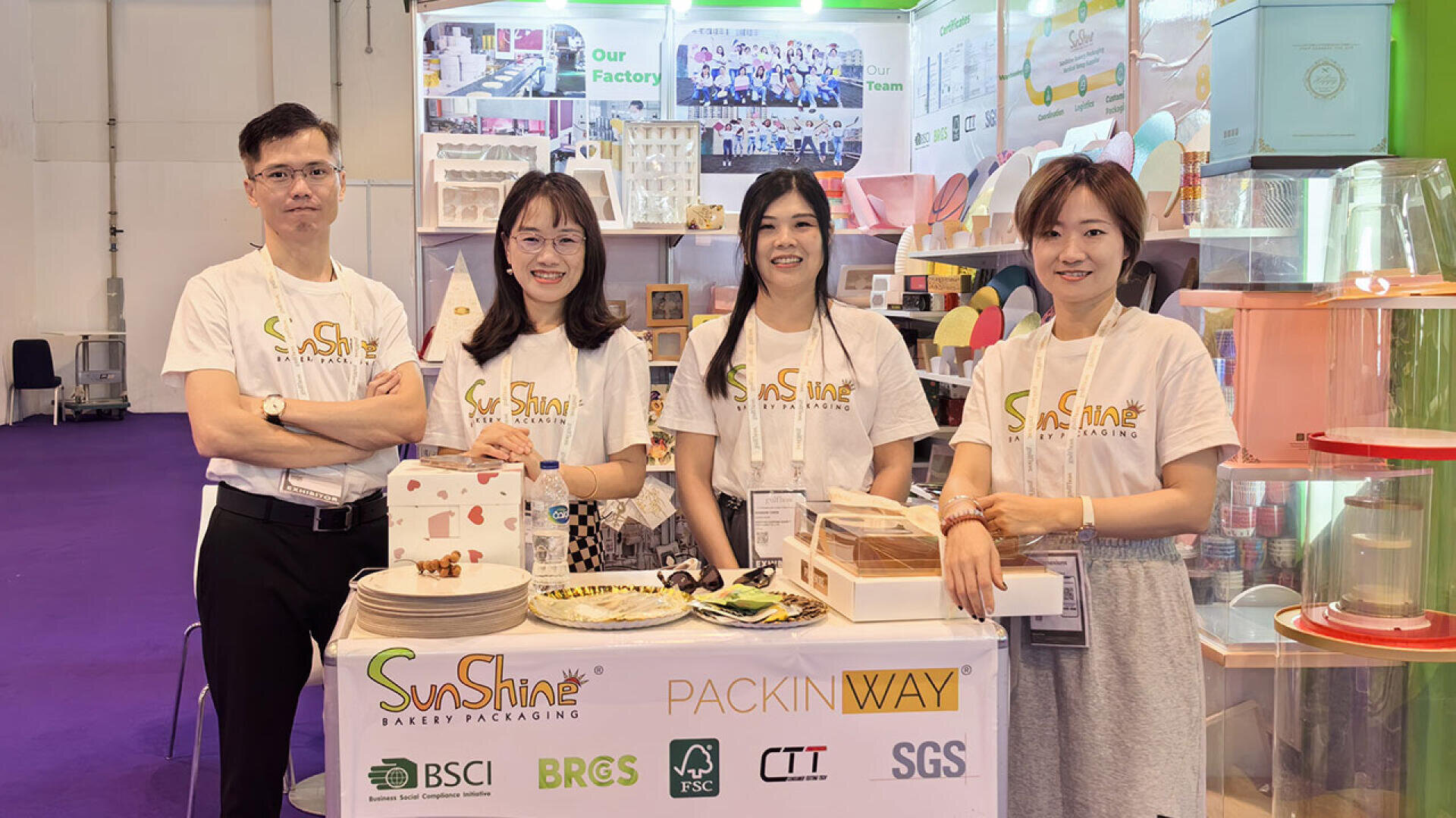 Four people wearing matching SunShine t-shirts stand and smile behind a bakery packaging booth with products and samples displayed on the table at a trade show or exhibition.