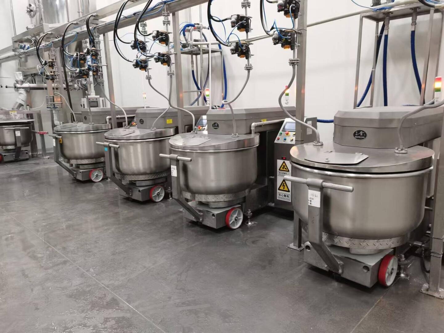 Four large industrial mixers with stainless steel bowls are lined up in a clean food processing facility, connected to overhead pipes and control panels, set on a polished concrete floor.