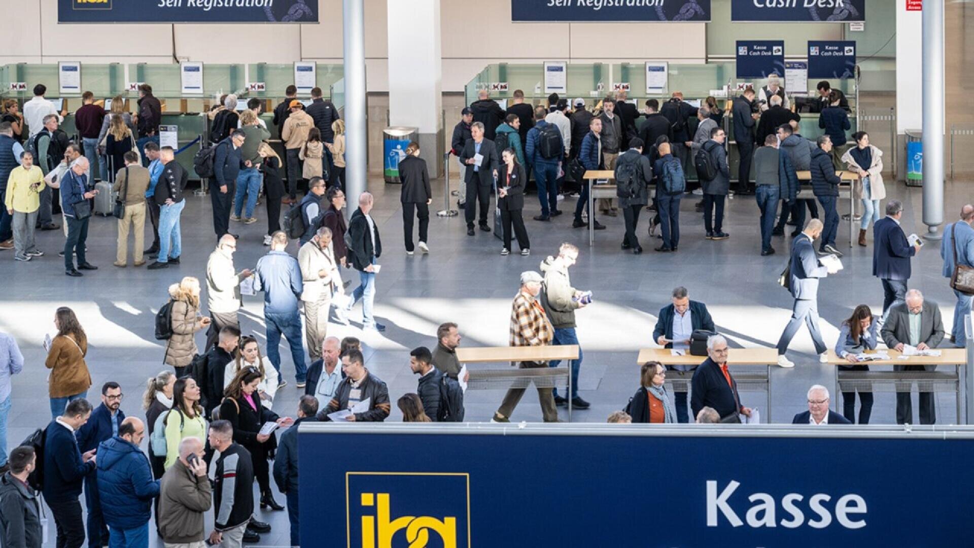 A busy registration area at a trade show or conference, with many people standing in lines, talking, and checking in at counters. A large blue sign with “Kasse” is visible in the foreground.