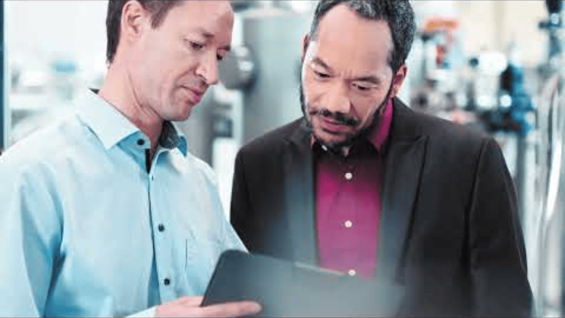 Two men standing indoors and looking at a tablet together; one is wearing a light blue shirt, and the other is in a dark blazer with a red shirt. They appear to be discussing or reviewing information.