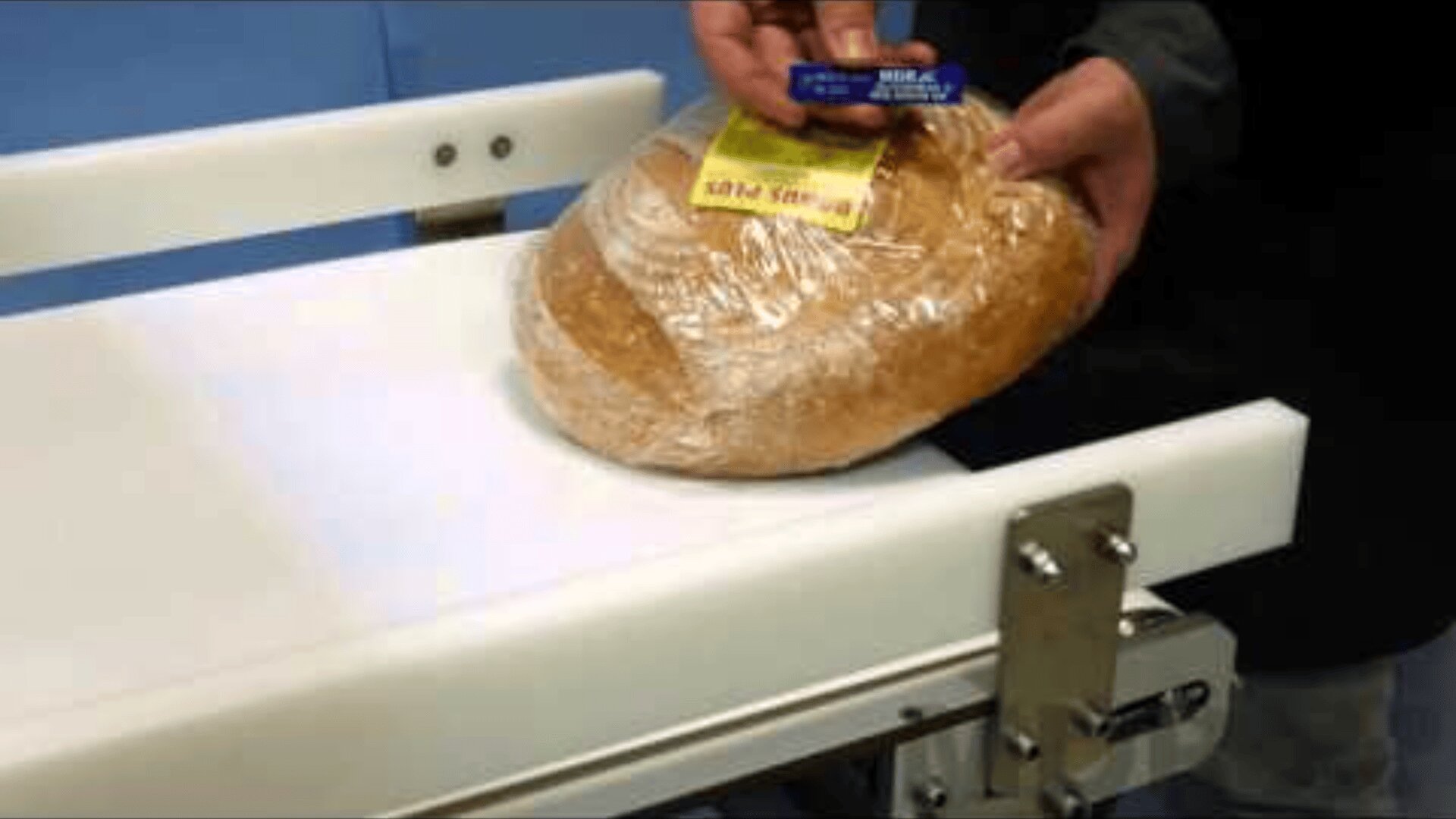 A person holds a round, plastic-wrapped loaf of bread on a white conveyor belt and prepares to scan the yellow price label with a small handheld scanner.