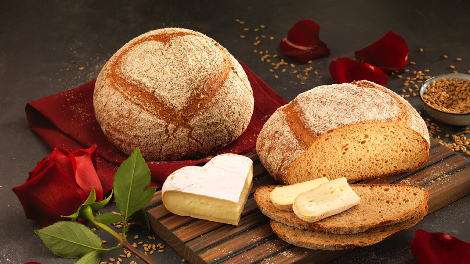 Two round loaves of rustic bread, one sliced with pieces of cheese, sit on a wooden board with a red napkin. Red rose petals and a whole rose are scattered around, creating a romantic atmosphere.
