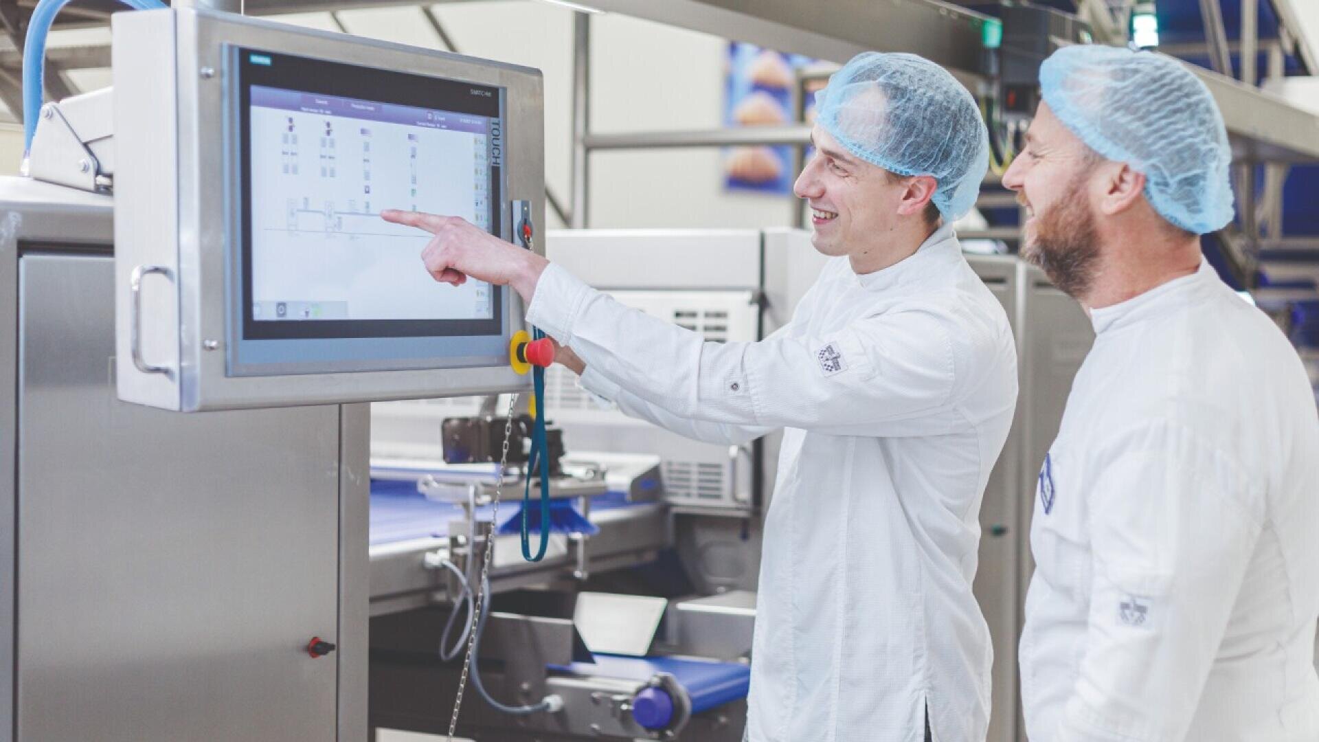 Zwei Personen in weißen Laborkitteln und Haarnetzen stehen neben industriellen Geräten, wobei eine von ihnen auf Daten zeigt, die auf einem großen Touchscreen-Monitor in einer sauberen, modernen Produktionsumgebung angezeigt werden.