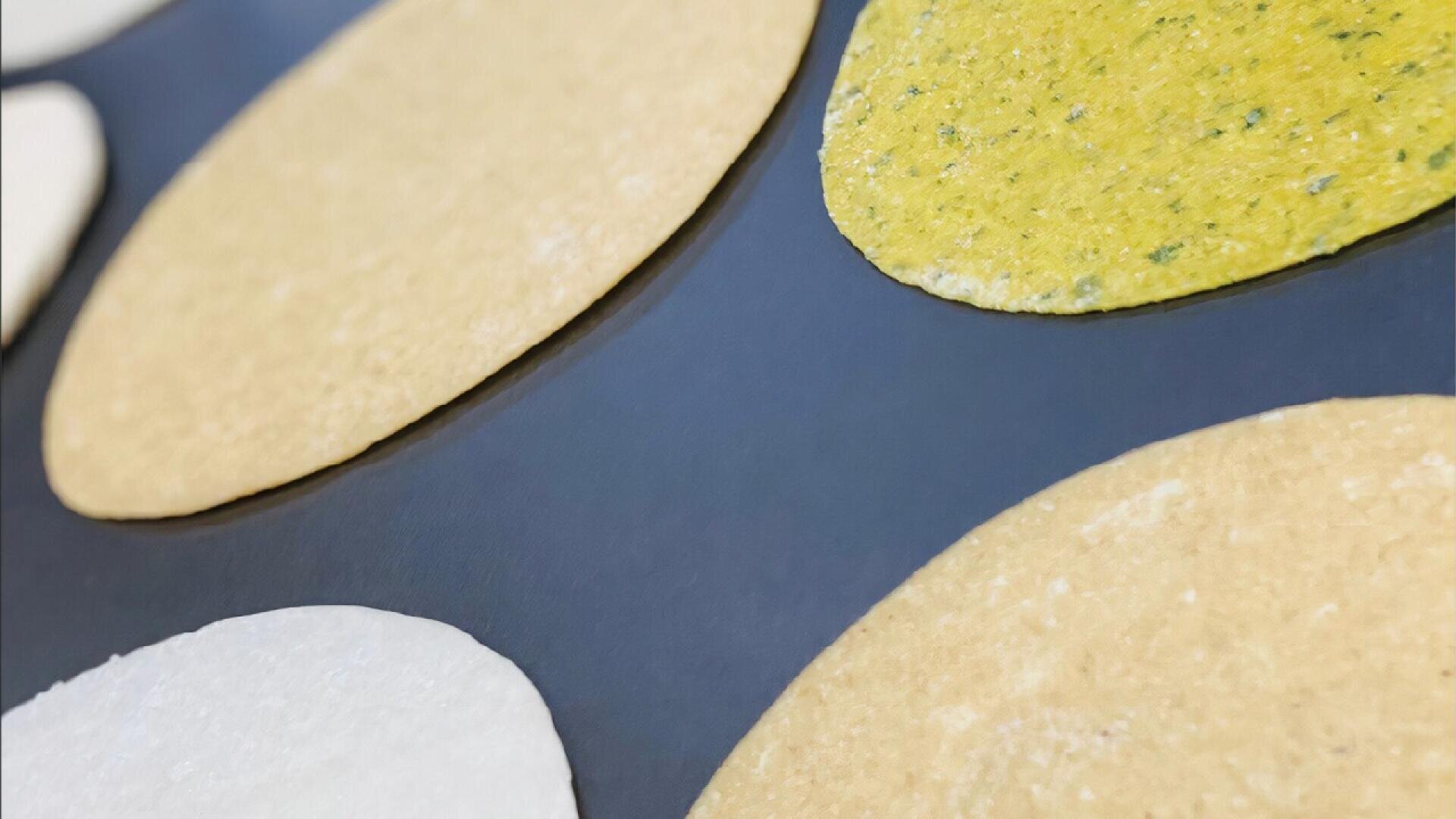 Nahaufnahme mehrerer ungekochter Tortillakreise in verschiedenen Farben - weiß, gelb und grün -, die auf einer dunklen Fläche angeordnet sind.