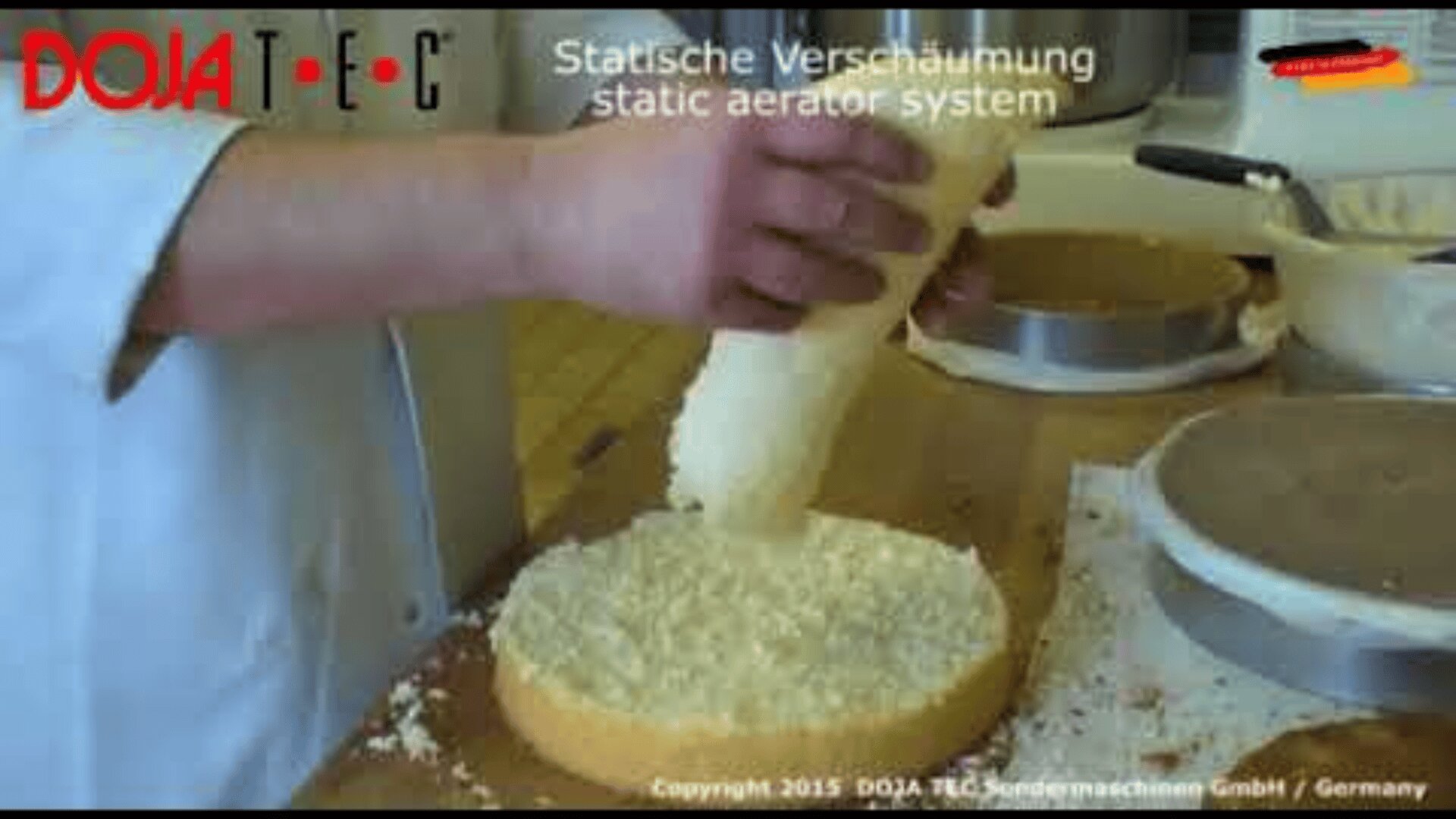 A person uses a cylindrical tool to apply a foamy substance onto a round cake base in a kitchen setting. The text Statische Verschäumung static aerator system appears at the top.