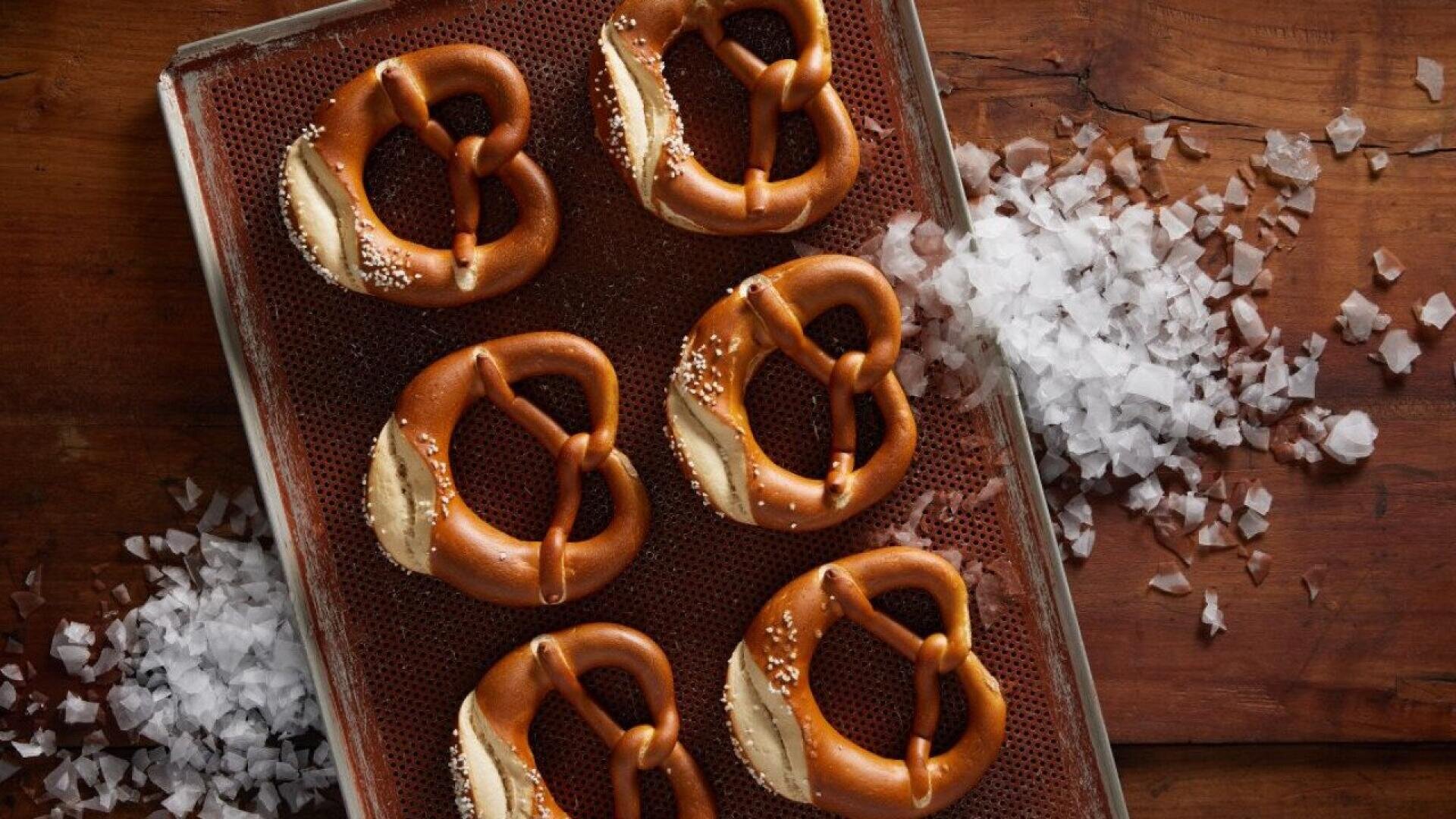 Six golden-brown pretzels sprinkled with coarse salt rest on a baking tray. Flakes of salt are scattered around the tray on a wooden surface.