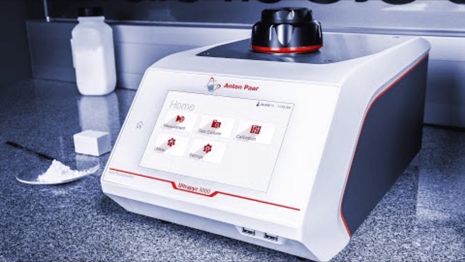 On a work surface is a laboratory instrument, the Anton Paar Density 5000. A touchscreen display shows menu options. Powder, a white bottle, and a cube are placed next to it, indicating sample analysis.