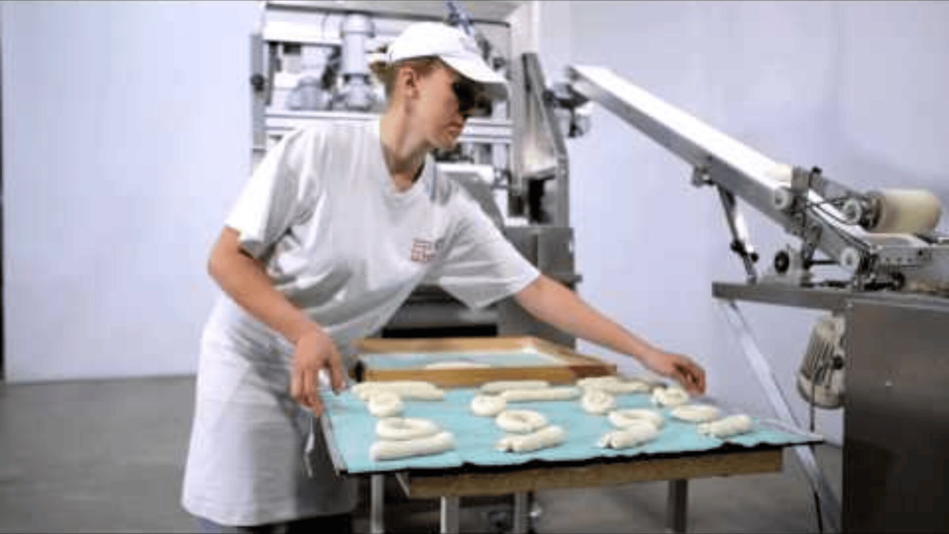 A person in a white uniform and cap places a tray of unbaked pretzels on a table next to industrial baking equipment in a commercial kitchen.