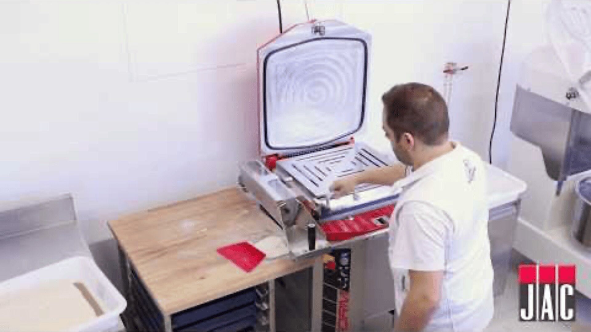 Ein Mann in einem weißen Hemd bedient eine Teigpressmaschine auf einem Holztisch in einem weißen Raum. Das JAIC-Logo ist in der unteren rechten Ecke zu sehen.